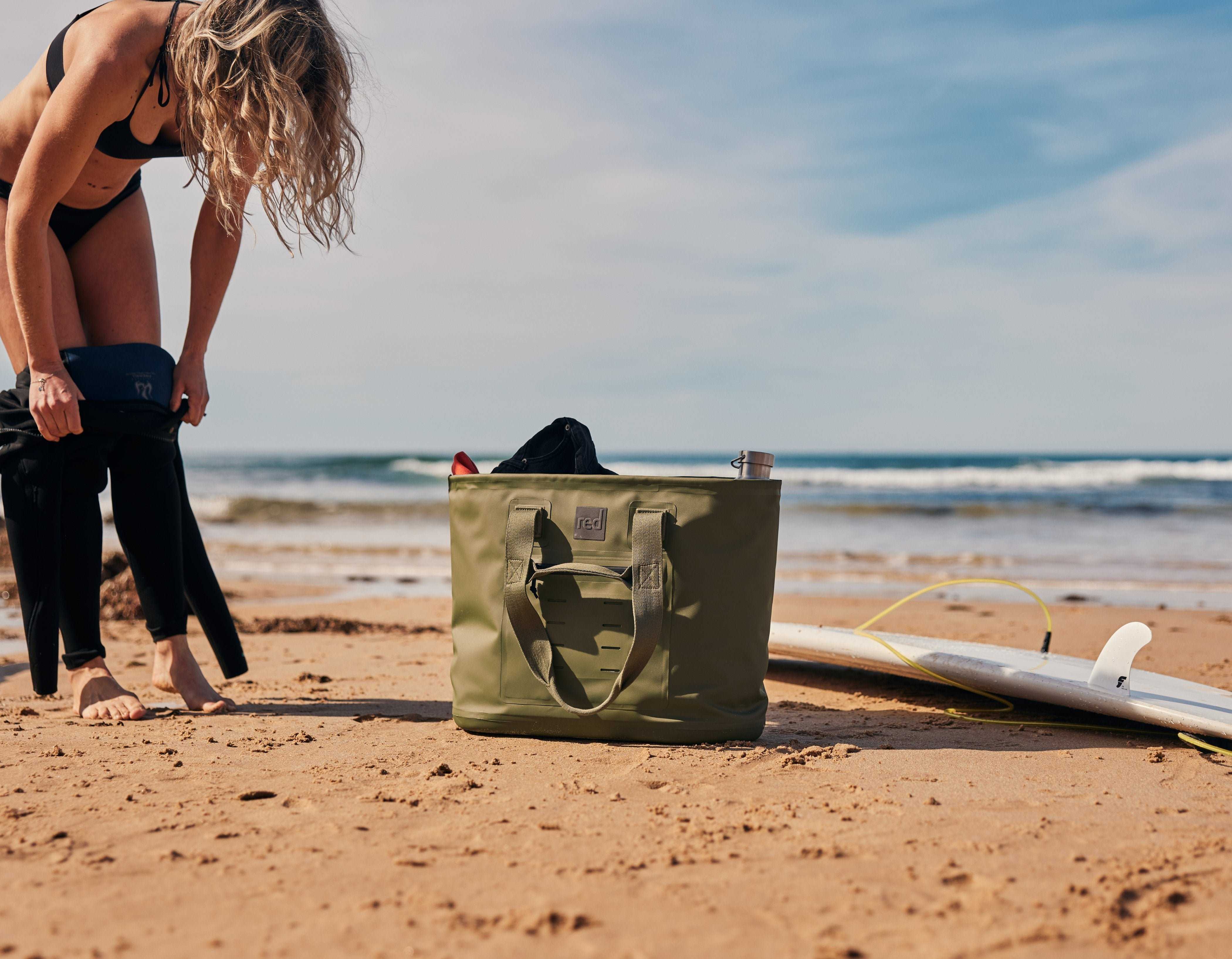 Woman changing on the beach beside a surf board and a Red Olive Green Waterproof Tote Bag.