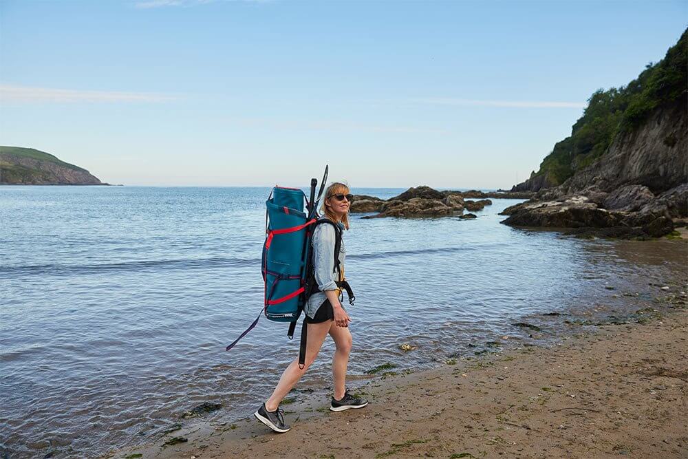 woman walking along a beach carrying a paddle board bag