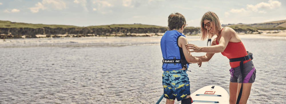 A woman helping her son put on his life jacket before getting on a paddleboard