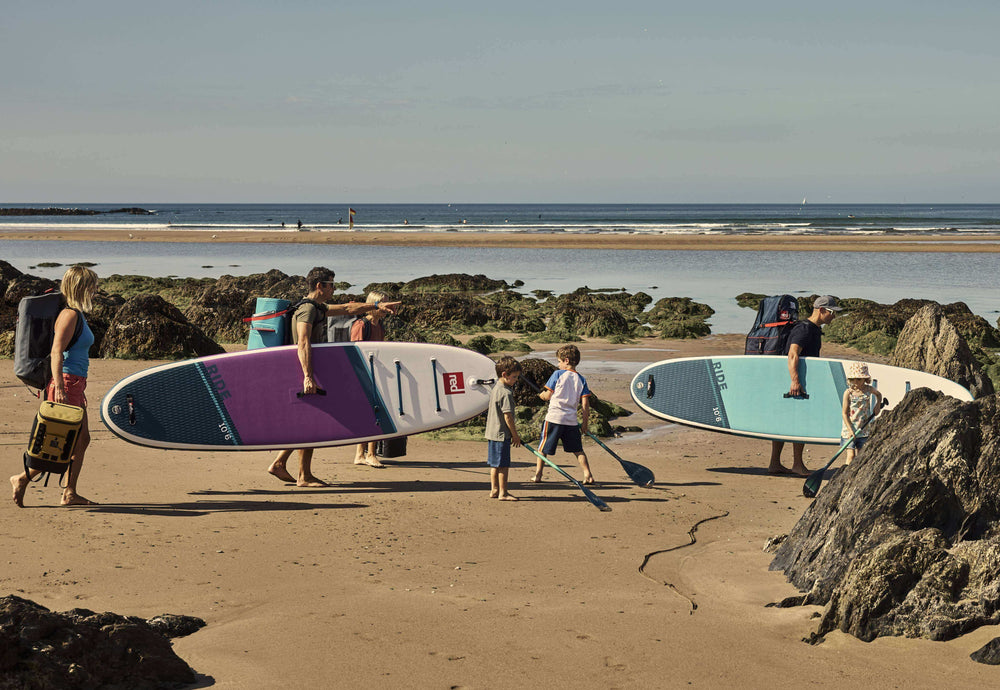 family walking on beach, carrying Ride paddle boards, cooler backpacks, kit bag, and paddles.