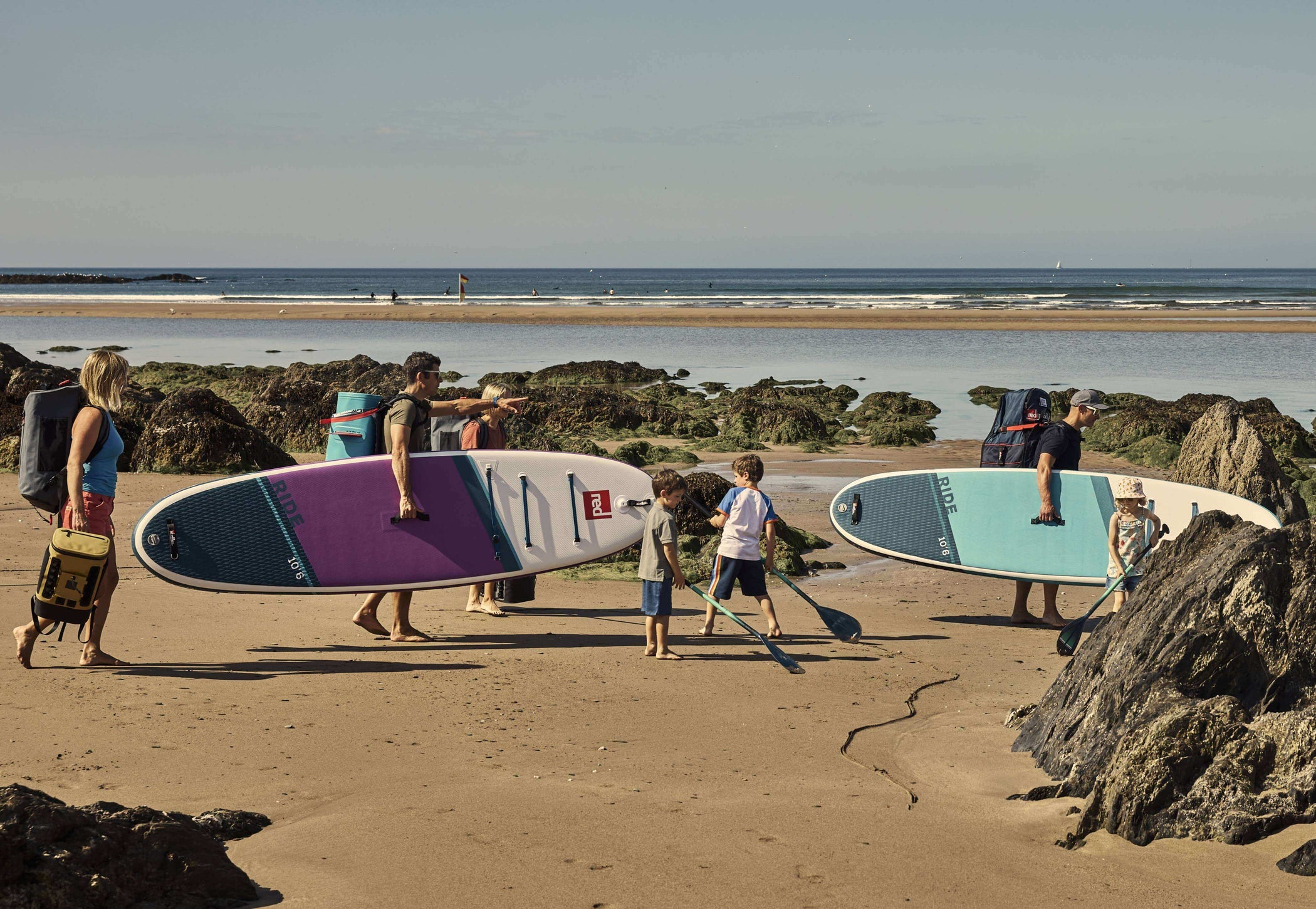 family walking on beach, carrying Ride paddle boards, cooler backpacks, kit bag, and paddles.