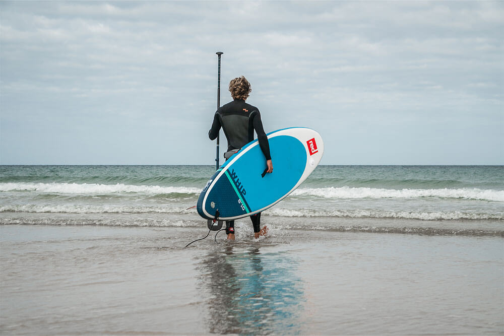 man wearing wetsuit walking into the ocean carrying Whip MSL inflatable paddle board