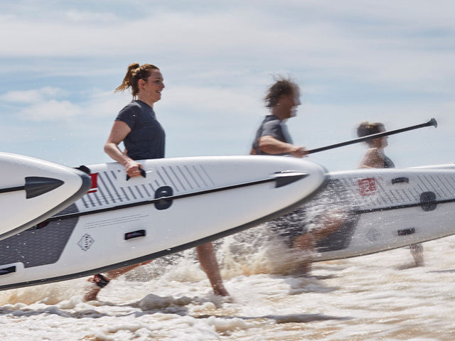 4 people running toward the sea carrying Red Paddle Co Racing Paddleboards