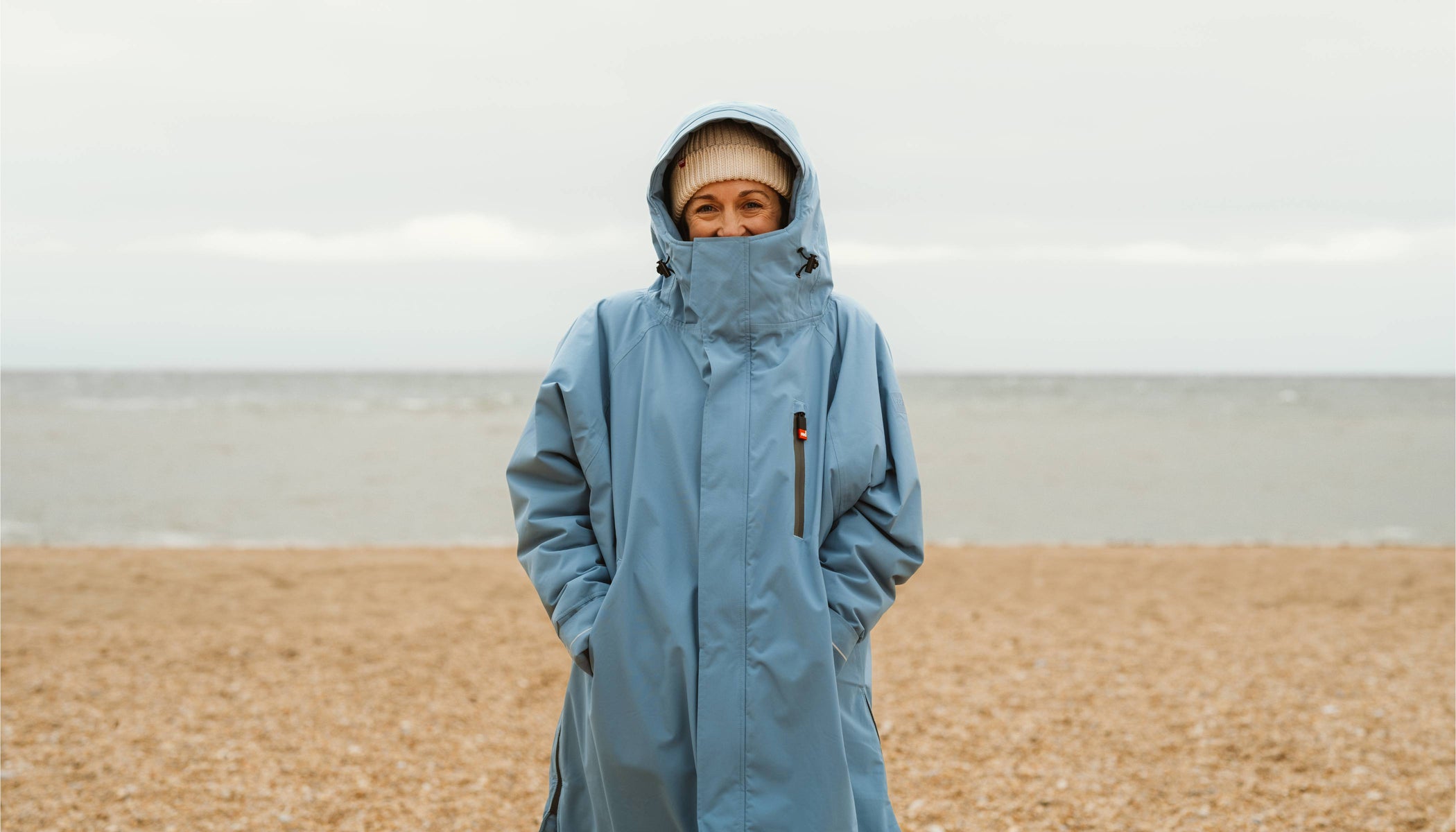Woman standing on beach, wearing a Red Equipment EVO Pro Changing Robe and Beanie with the sea behind her.