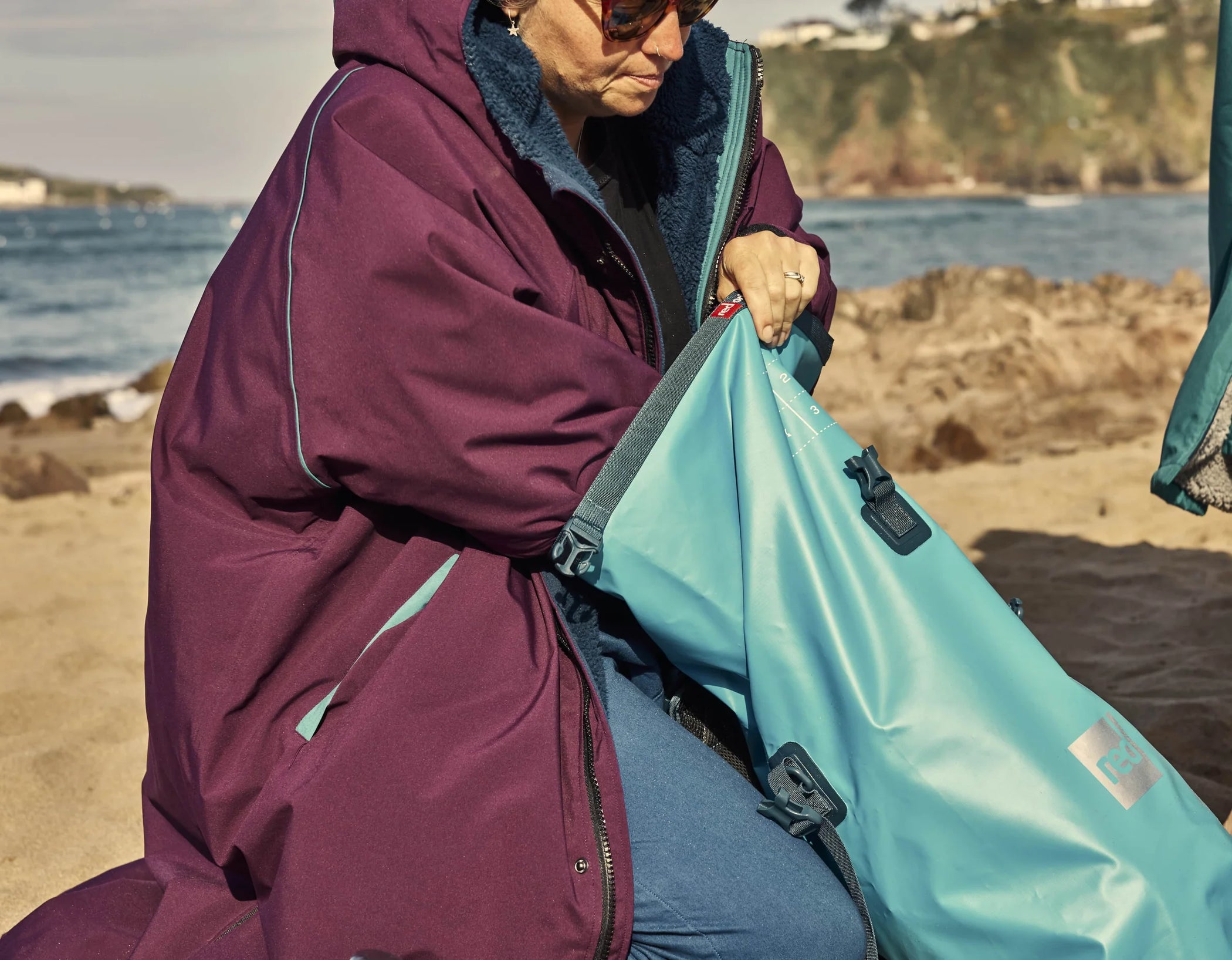 A woman placing things into her blue stash bag whilst sat on the beach