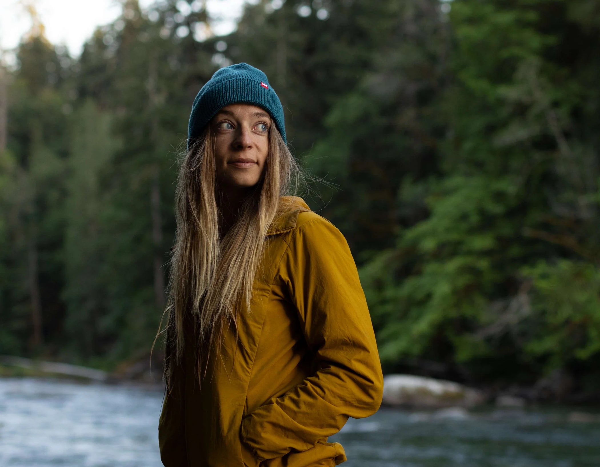 A woman wearing the eddy beanie, in the woods standing in front of a river looking into the distance