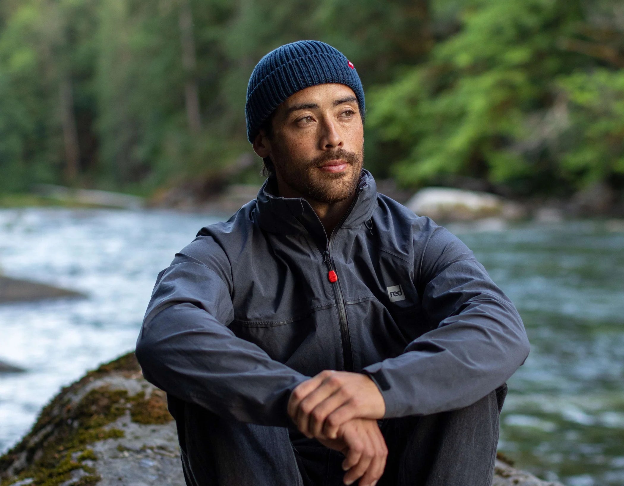 A man sat on a rock by a river wearing the flow beanie