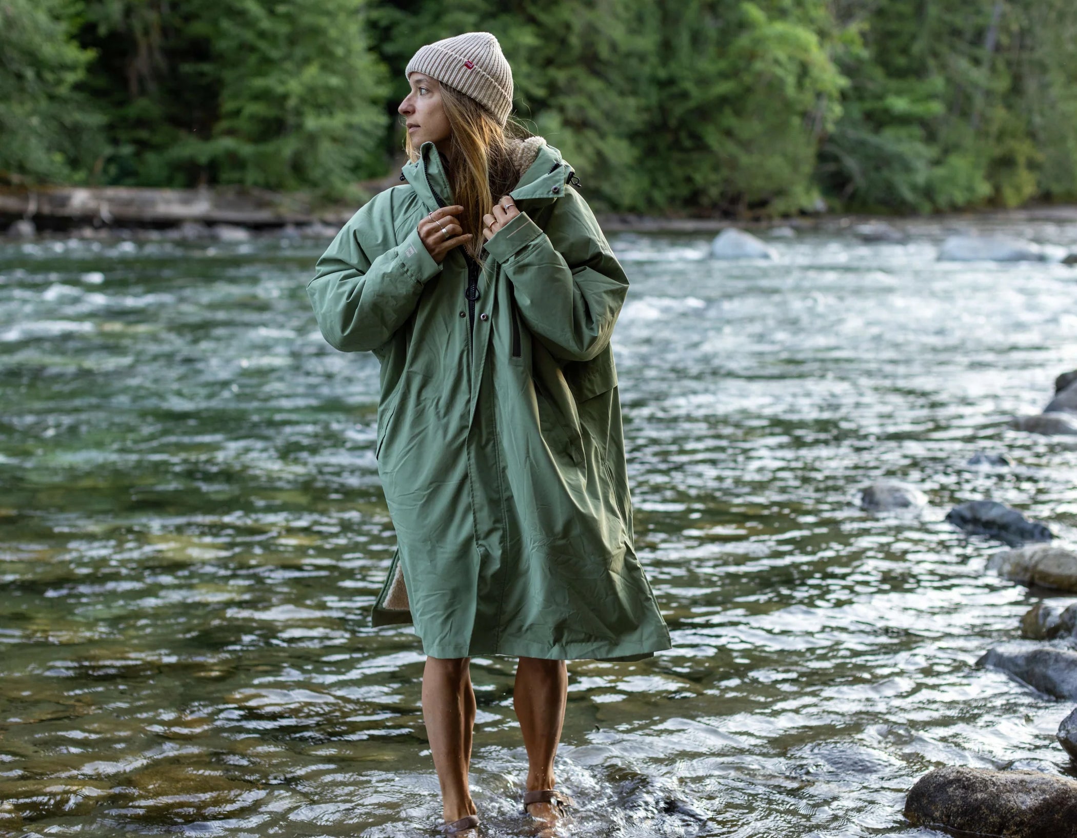 A woman wearing her long sleeve changing robe and flow beanie, paddling in a river, looking to the side