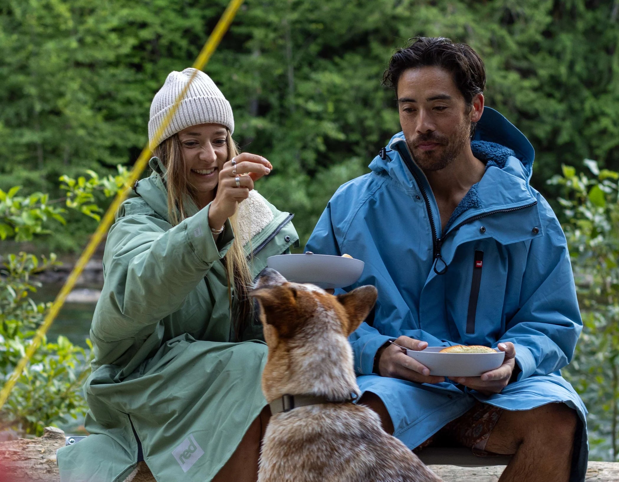 A man and woman sat down eating and feeding their dog, wearing their long sleeve changing robes