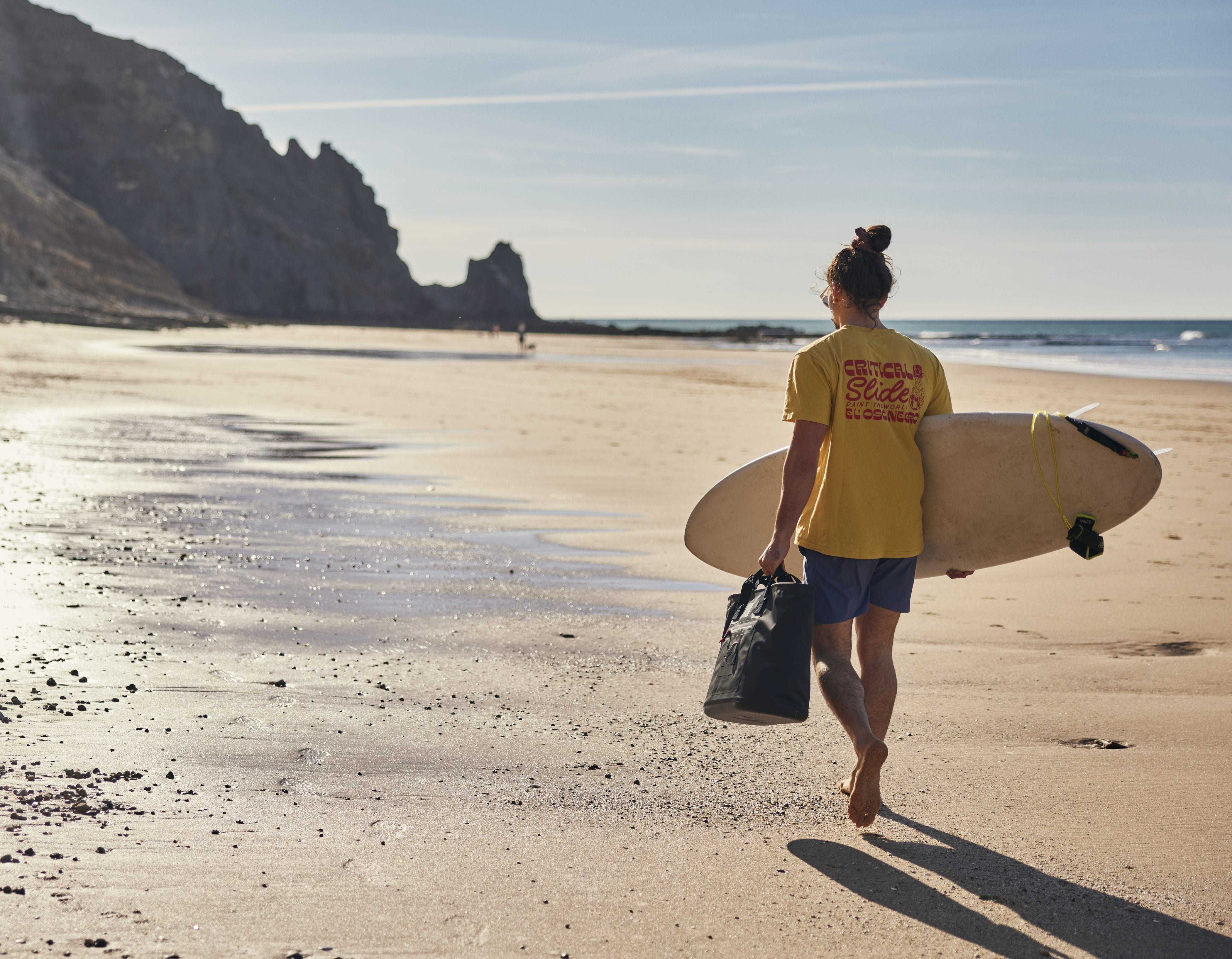 Man carrying Red's Waterproof Tote bag and surf board across the beach.